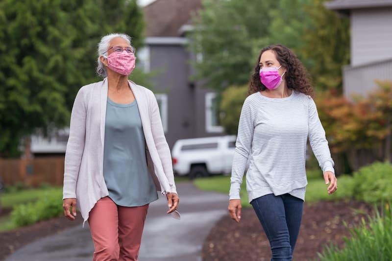 A-mixed-race-senior-adult-woman-is-out-for-a-walk-with-her-adult-daughter.-The-two-women-are-wearing-protective-face-masks